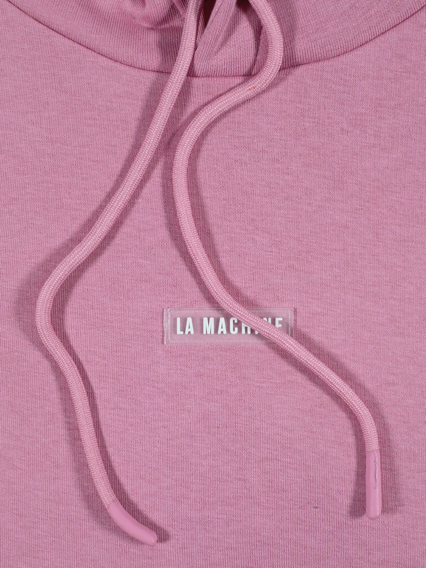 Close-up of a pink Vai Allez Vamos hoodie by La Machine Cycle Club featuring matching drawstrings and a small chest patch with blurred white LA MACHINE text—ideal for cycling apparel fans.