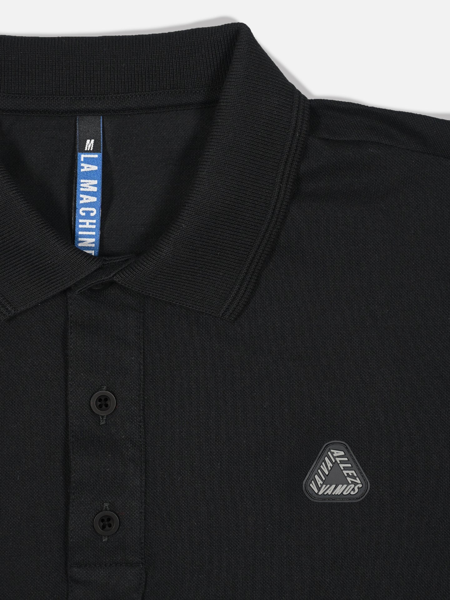 Close-up of a black Vai Allez Vamos polo shirt with collar, two buttons, and a Viva Allezz Manus chest badge. The neck tag shows La Machine Cycle Club, size M—ideal for cycling fans.