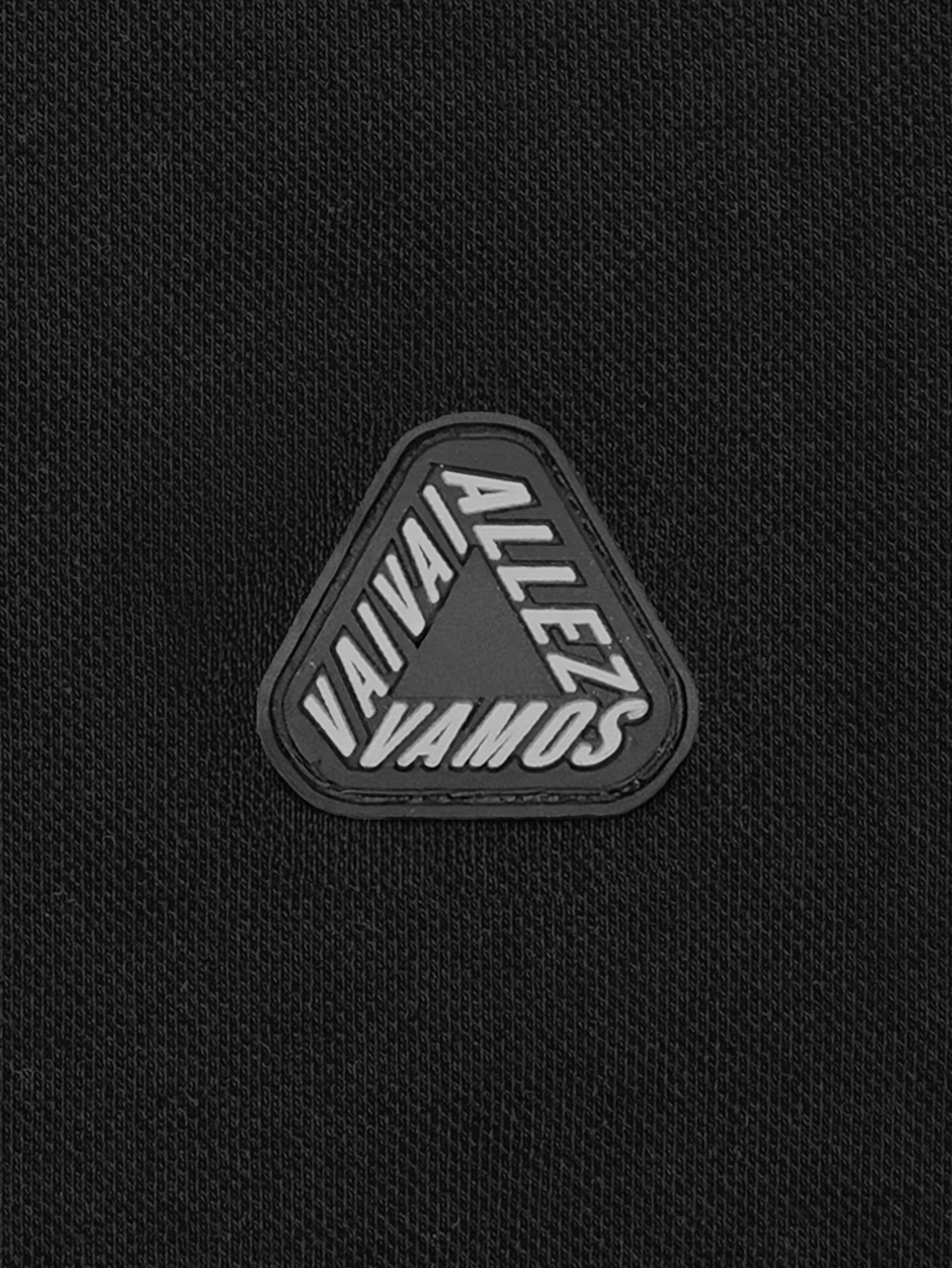 The Vai Allez Vamos - Polo Shirt - Black by La Machine Cycle Club features black textured cycling fabric with a triangular black patch at the center, displaying VAVÁ!, ALLEZ!, and VAMOS in white on each side for a unisex look.