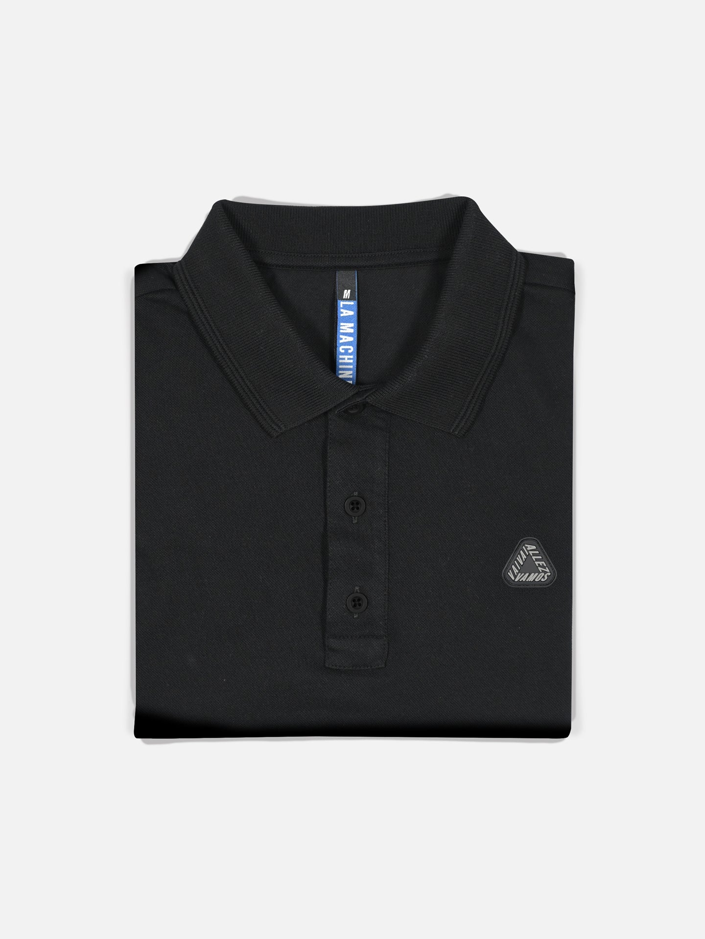 The Vai Allez Vamos polo shirt by La Machine Cycle Club is a black unisex style with a collar, three buttons, and a small embroidered logo on the chest, shown neatly folded against a plain light background.