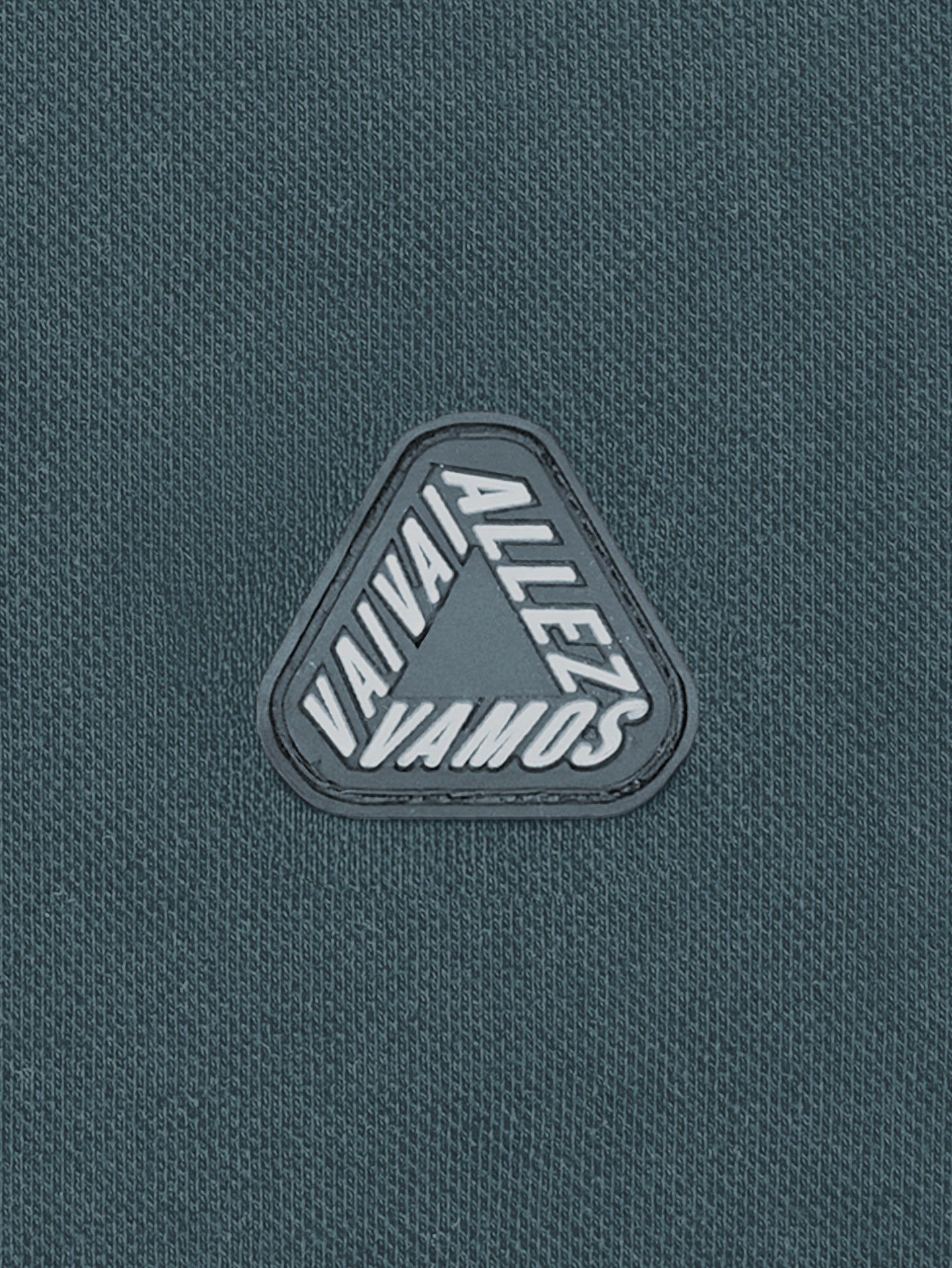A close-up of the La Machine Cycle Club Vai Allez Vamos unisex polo shirt in Orion Blue, featuring a triangular patch with the Tour de France cheers: VAVAT, ALLEZ, and VAMOS in bold white letters.