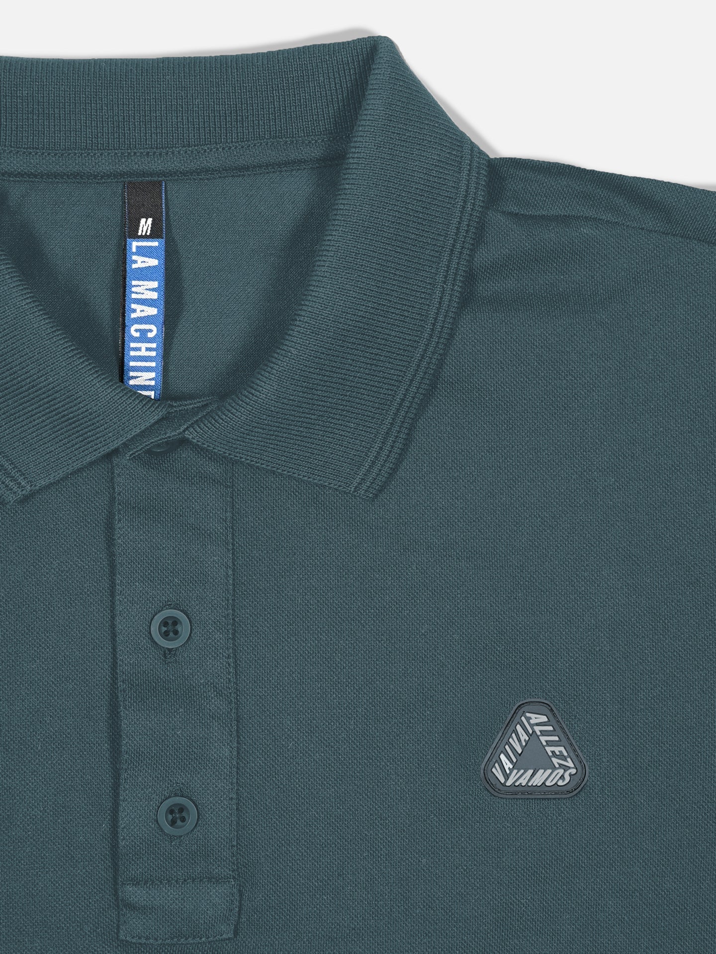 The La Machine Cycle Club Vai Allez Vamos Polo Shirt in Orion Blue is a unisex polo with a folded collar, two-button placket, and chest patch reading VAMOS ALLLLL VAMOS. Tag says LA MAQUINA, size M. Inspired by the Giro d’Italia.