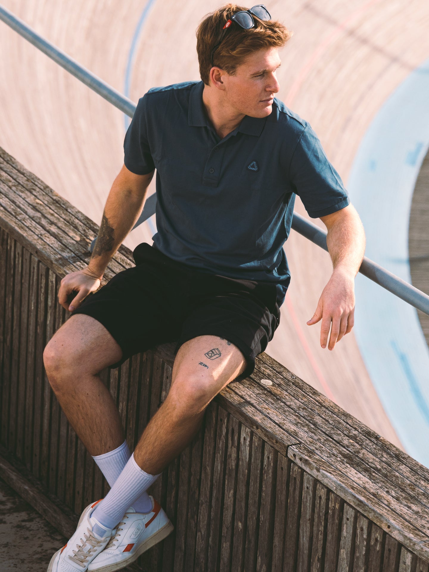 A young man with light brown hair sits on a wooden fence at an outdoor velodrome, wearing the La Machine Cycle Club Vai Allez Vamos Polo Shirt in Orion Blue, styled with black shorts and white sneakers as he evokes the spirit of the Giro d’Italia.