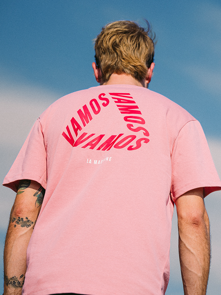 A blond person faces away, wearing La Machine Cycle Club’s Vamos - Loose Fit - Vuelta a España T-shirt, featuring bold red VAMOS prints on the pink back. Their tattooed arm hints at a passion for Spanish cycling. The sky is blue.