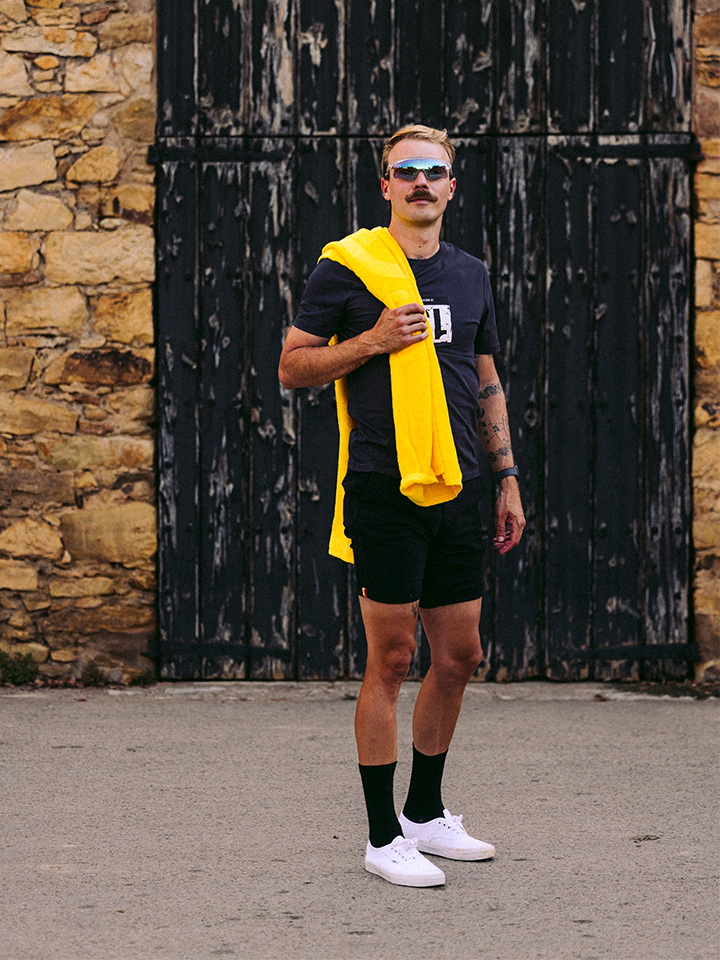 A man with a mustache stands before a rustic stone wall and black doors, wearing sunglasses, dark clothes, and has the Le Tour de la Plage Beach Towel by La Machine Cycle Club draped over his shoulder.