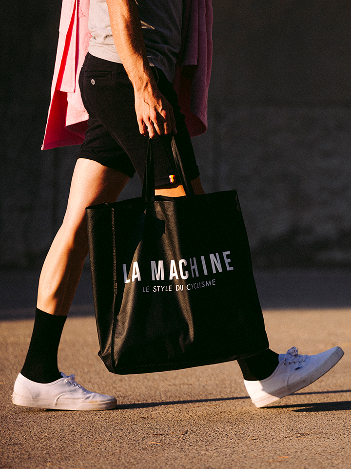 A person walks outdoors carrying the La Machine - Shopper by La Machine Cycle Club, a large black non-woven bag featuring the brand’s logo and “LE STYLE DU CYCLISME,” paired with black shorts, black socks, white sneakers, and a pink shirt over their shoulder.