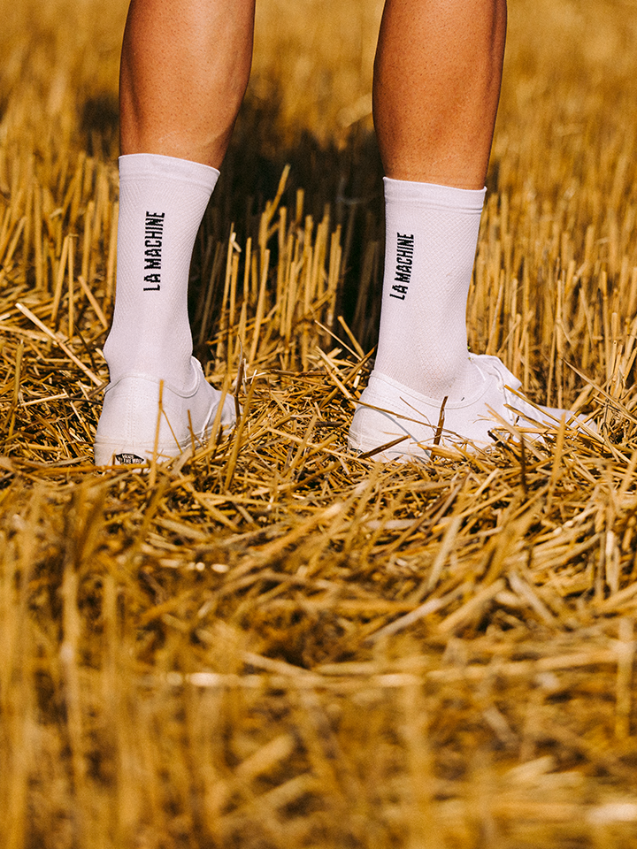 A person stands on dry, golden straw, wearing white La Machine Logo Cycling Socks by La Machine Cycle Club and white sneakers. Only their lower legs and feet are visible.
