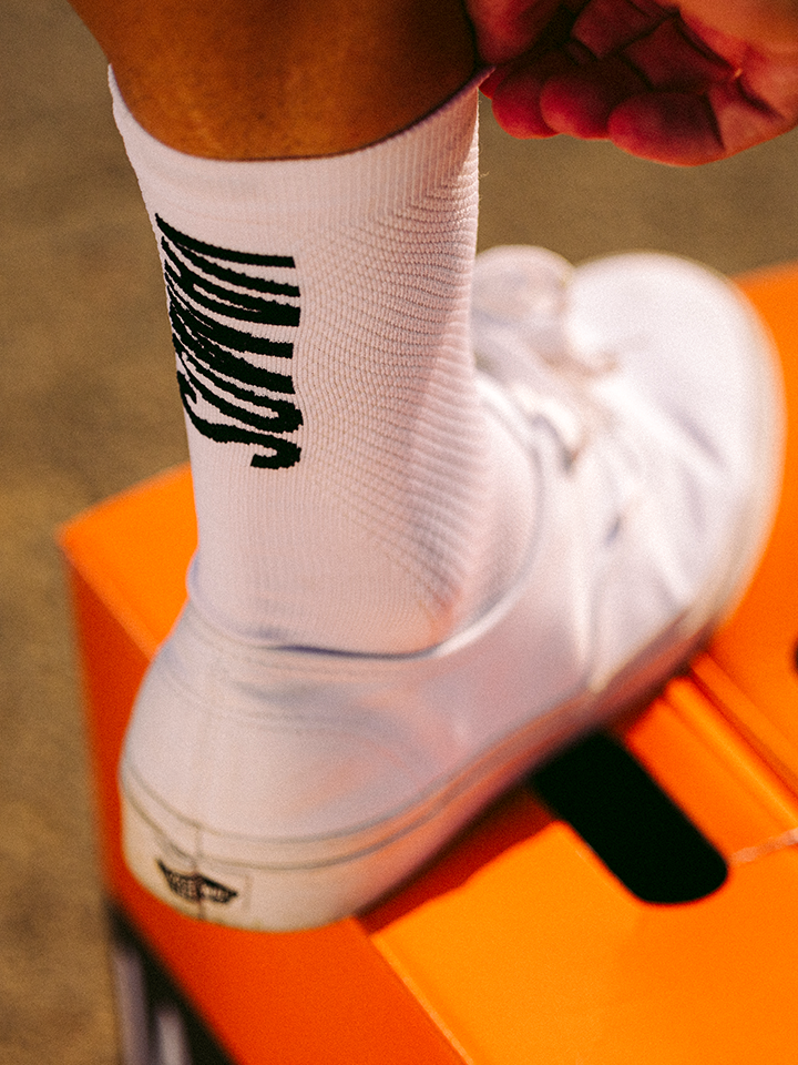 A person wearing Vamos - Cycling Socks by La Machine Cycle Club, featuring all-white fabric, bold black lettering, and optimum elastic fit, pulls up a sock while standing on an orange box.