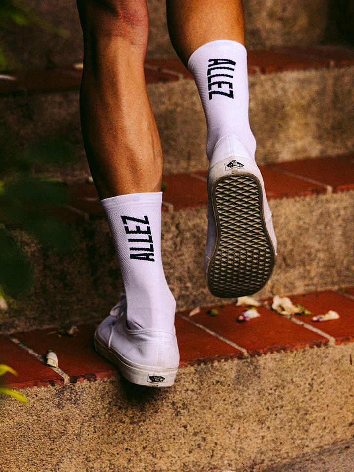 A person wears La Machine Cycle Club’s Allez Allez – Cycling Socks in all white, featuring a seamless toe, paired with white sneakers while walking up outdoor brick steps. Only their legs and feet are visible.
