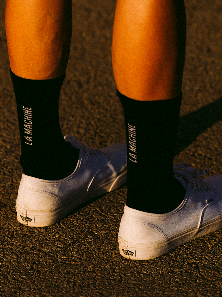 A person wearing white sneakers and La Machine Cycle Clubs black La Machine Logo Cycling Socks (merino), standing on asphalt in warm sunlight.