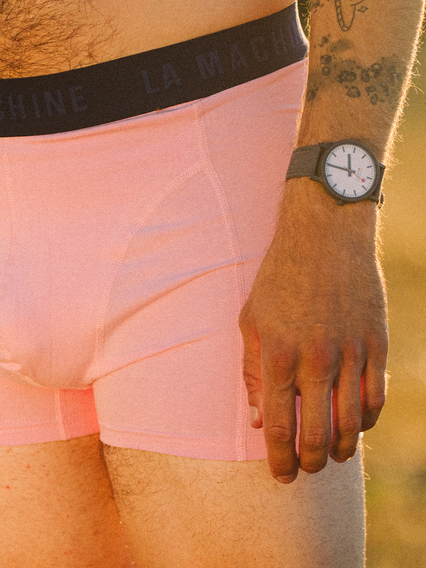 A person stands outdoors, showing only their lower torso and one arm. Theyre wearing La Machine Cycle Club’s Giro Pink boxershorts with LA MACHINE on the waistband, and a gray wristwatch is on their wrist.