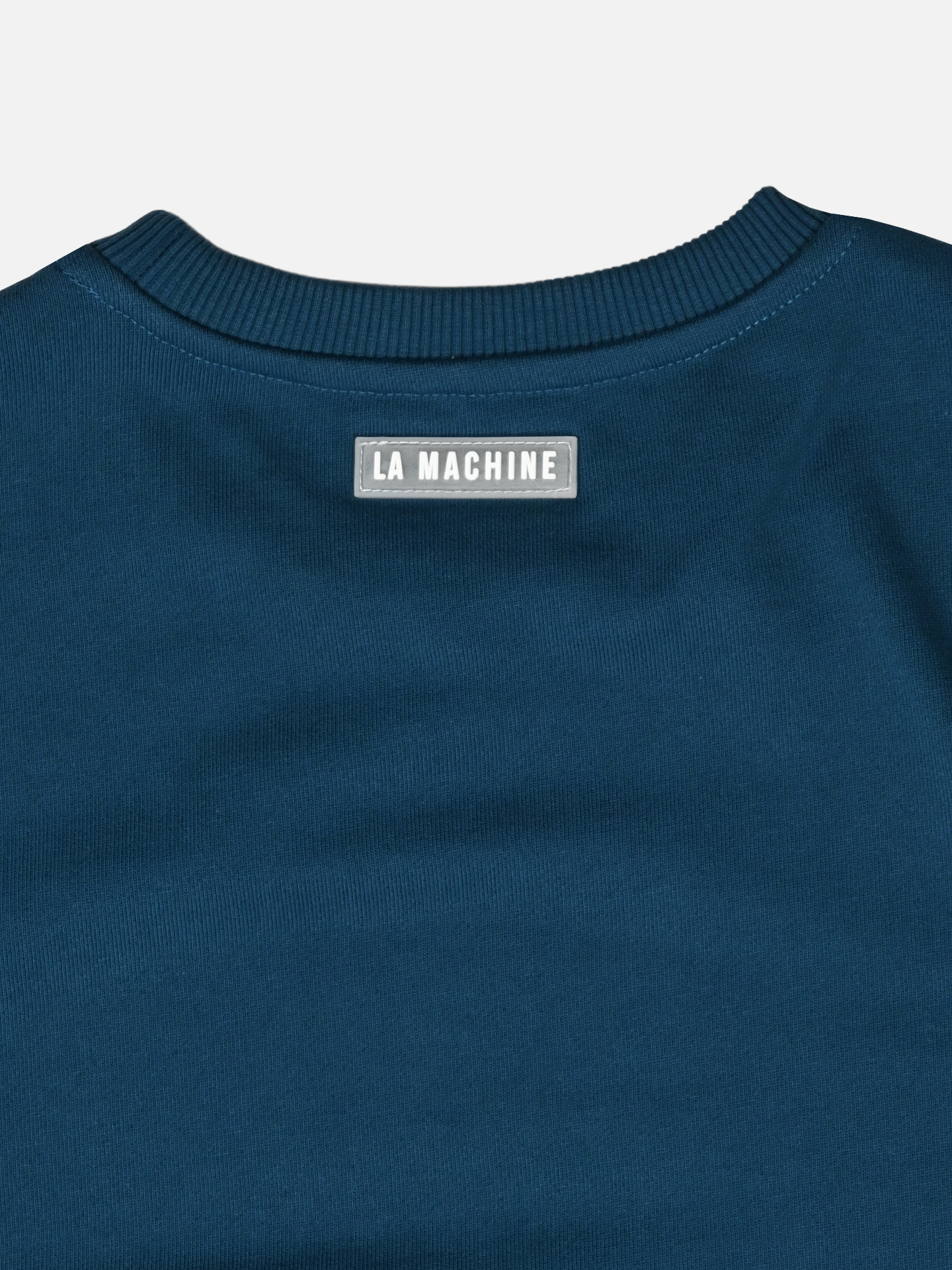 Close-up of the upper back of the Coffee Ride Winners Club - Sweatshirt in Sailor Blue by La Machine Cycle Club, featuring a rectangular label with LA MACHINE stitched below the ribbed collar—ideal for your next coffee ride.