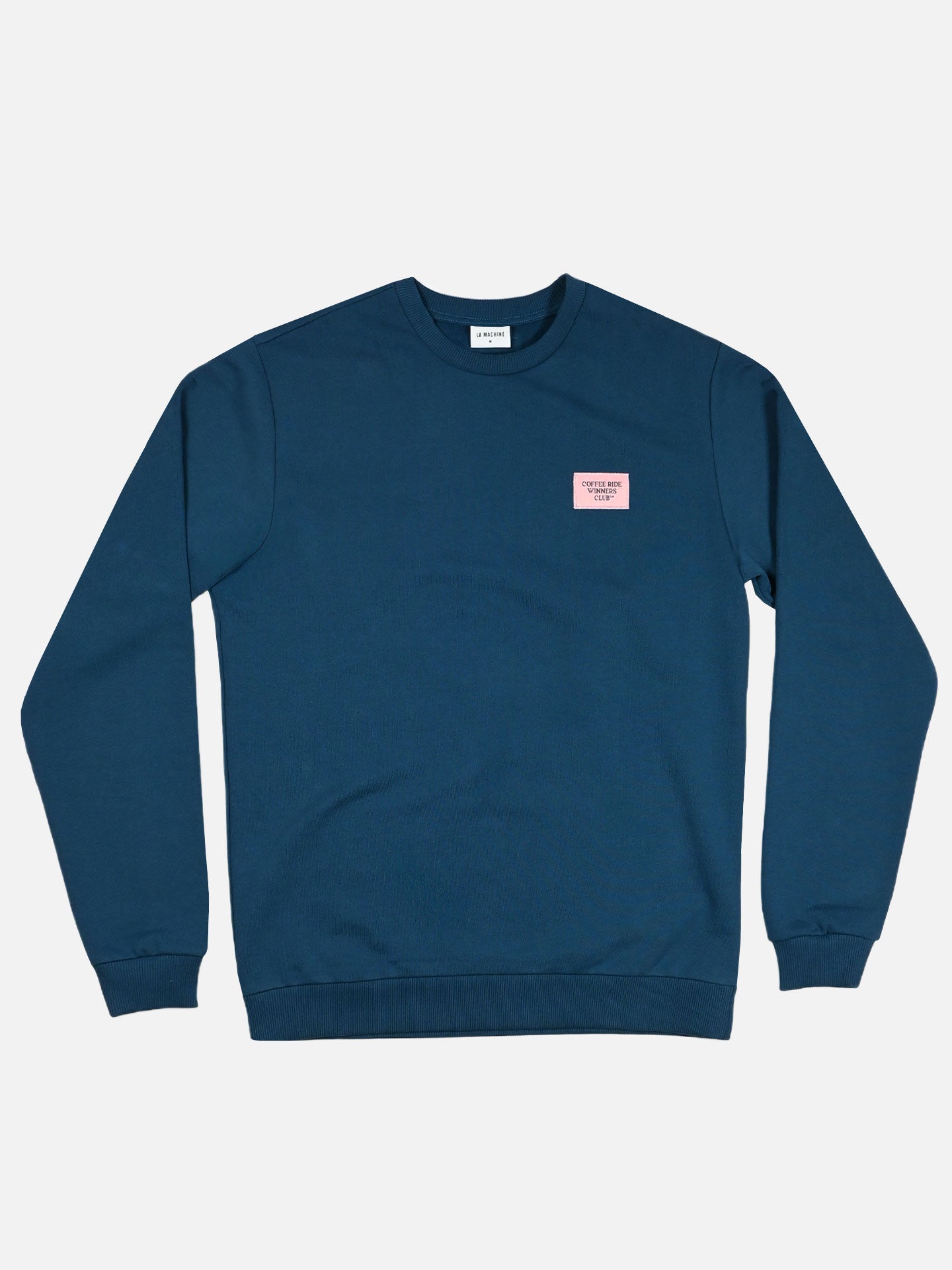 The Coffee Ride Winners Club Sweatshirt by La Machine Cycle Club features a navy blue design with a small pink “GOOD THINGS TAKE TIME” patch, crew neckline, and ribbed cuffs—ideal for coffee rides or post-cycling style.