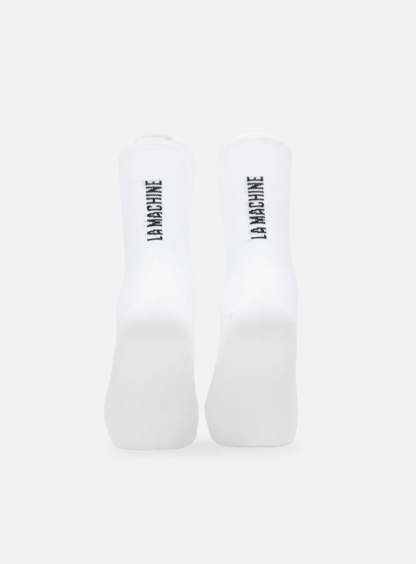 A pair of La Machine Logo - Cycling Socks in white by La Machine Cycle Club shown vertically on a plain light grey background, with bold black LA MACHINE branding on the soles.