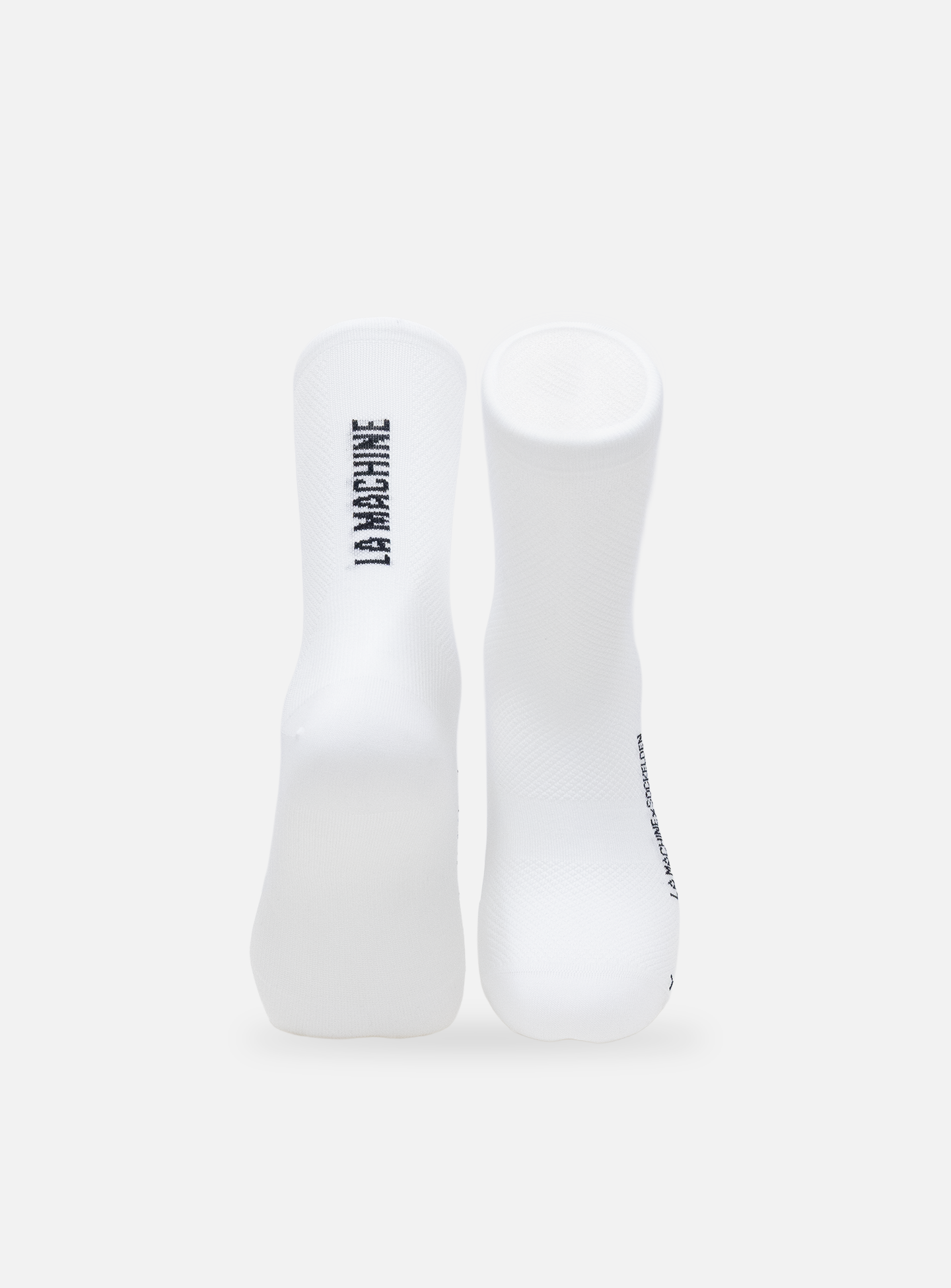 A pair of La Machine Logo - Cycling Socks - White by La Machine Cycle Club stand upright on a plain background, featuring “LA MACHINE” printed vertically in black on the left sock.