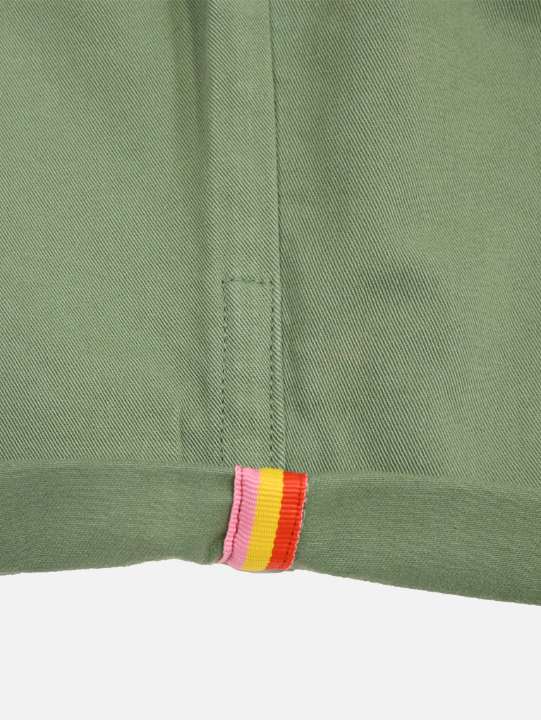 The Grand Tours - Rest Day Bermuda in Dusty Green by La Machine Cycle Club features sturdy cotton, a visible seam, folded hem, and a small vertical tab at the hem with pink, red, yellow, and white stripes inspired by classic Grand Tour jerseys.