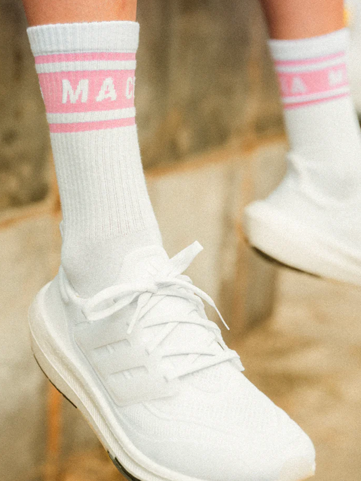 Close-up of a person wearing white athletic shoes and La Machine - Crew Socks - Giro Pink from La Machine Cycle Club. The socks feature partially visible text, with a blurred stone or concrete background.