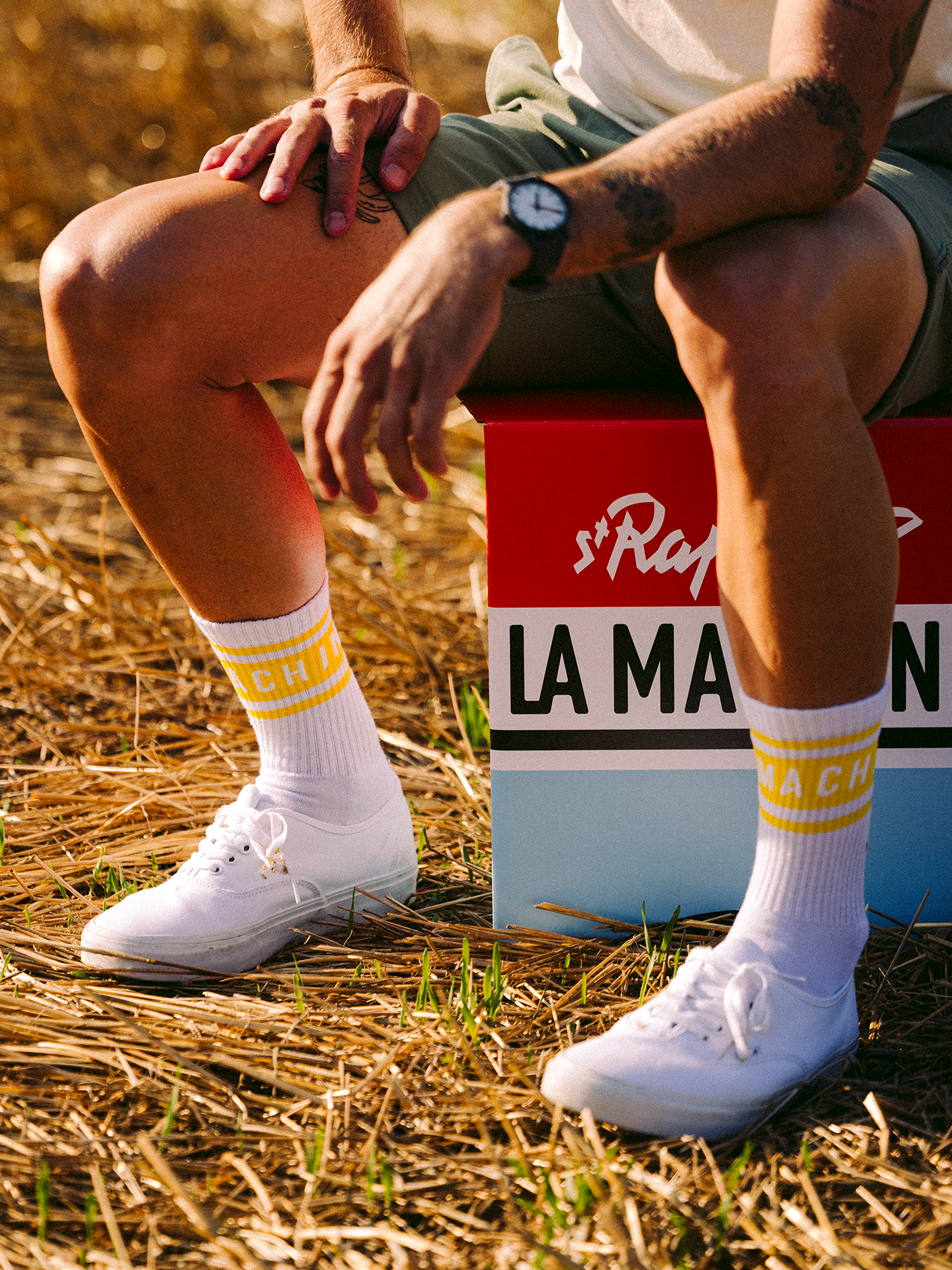 In a sunlit field, someone sits on a colorful LA MACHINE box, wearing La Machine Cycle Club’s La Machine - Crew Socks - Tour Yellow—white crew socks with Le Tour-yellow stripes and MACHINE text—plus white sneakers, shorts, and a watch. Only legs and hands show.