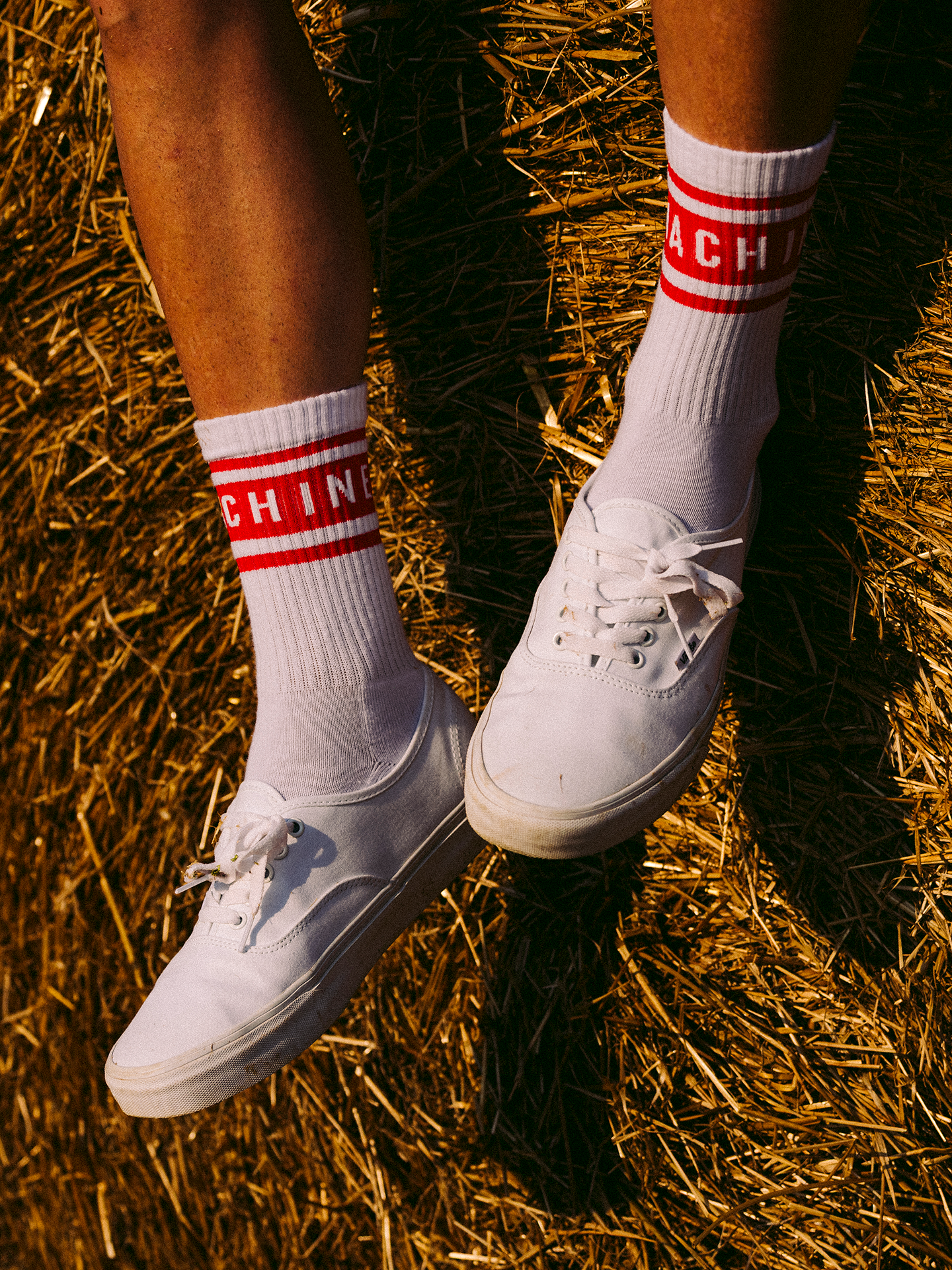 Wearing La Machine - Crew Socks - Vuelta Red from La Machine Cycle Club, a person sits on hay with legs crossed at the ankles. The white socks with red stripes and text evoke classic cycling style.