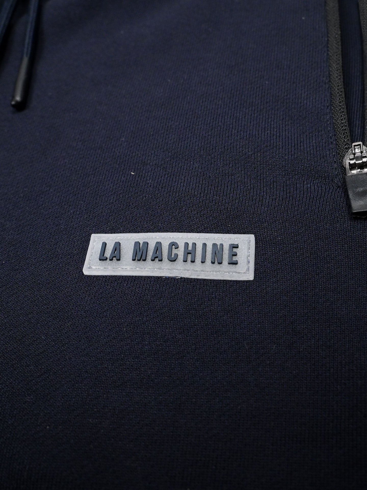 Close-up of dark navy Beyond the bike Sweatpants by La Machine Cycle Club, featuring a grey rectangular LA MACHINE patch in bold uppercase letters and a partial zipper on the right side.