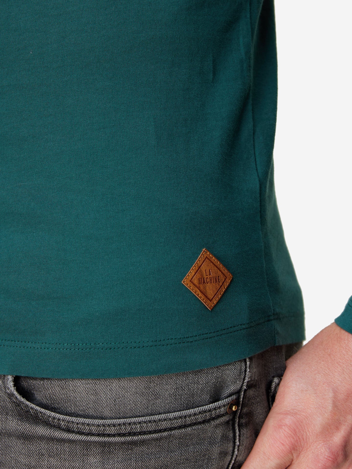 A close-up of a person wearing the Pedaleur de Charme long sleeve T-shirt by La Machine Cycle Club, featuring a small brown diamond-shaped La Machine patch, paired with gray jeans. Inspired by Hugo Koblet’s classic style.