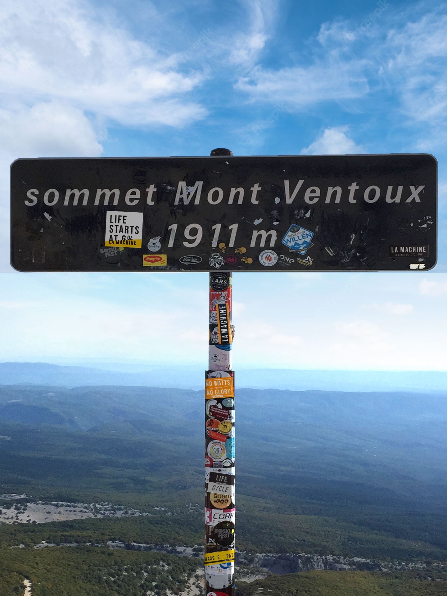 A sign at Mont Ventouxs summit, reading sommet Mont Ventoux 1911 m and adorned with La Machine Cycle Club’s Orange & Black Sticker Set (3x), stands against a scenic mountain and blue sky backdrop.
