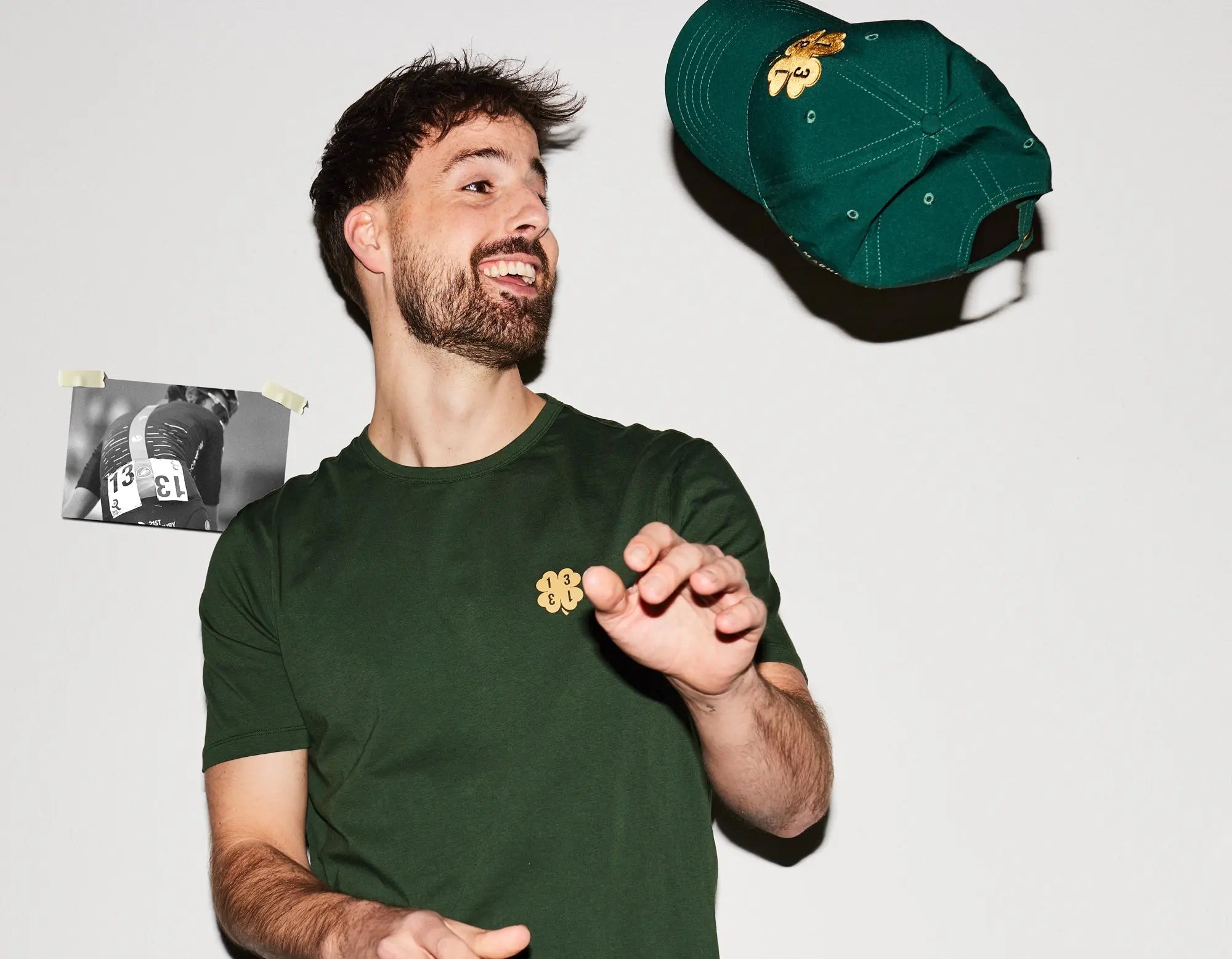 A smiling man in a green t-shirt tosses a matching green baseball cap in the air. A black-and-white photo is taped to the white wall behind him.