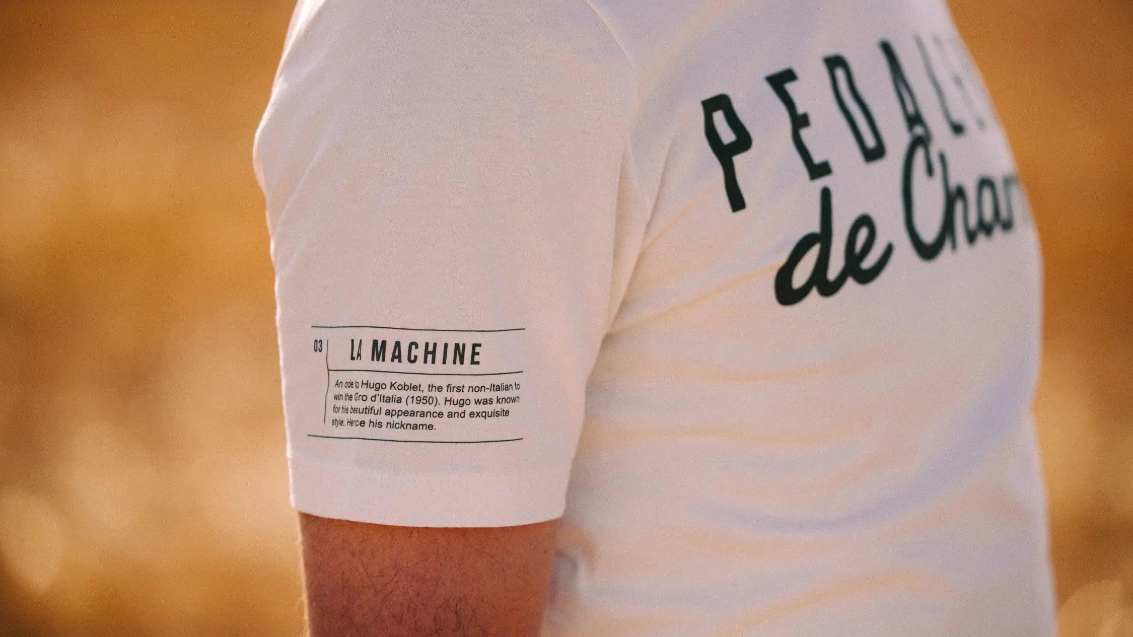 A person wearing a white T-shirt with PEDALEUR de Charme on the back and a LA MACHINE label with small text on the sleeve, standing outdoors in warm sunlight.