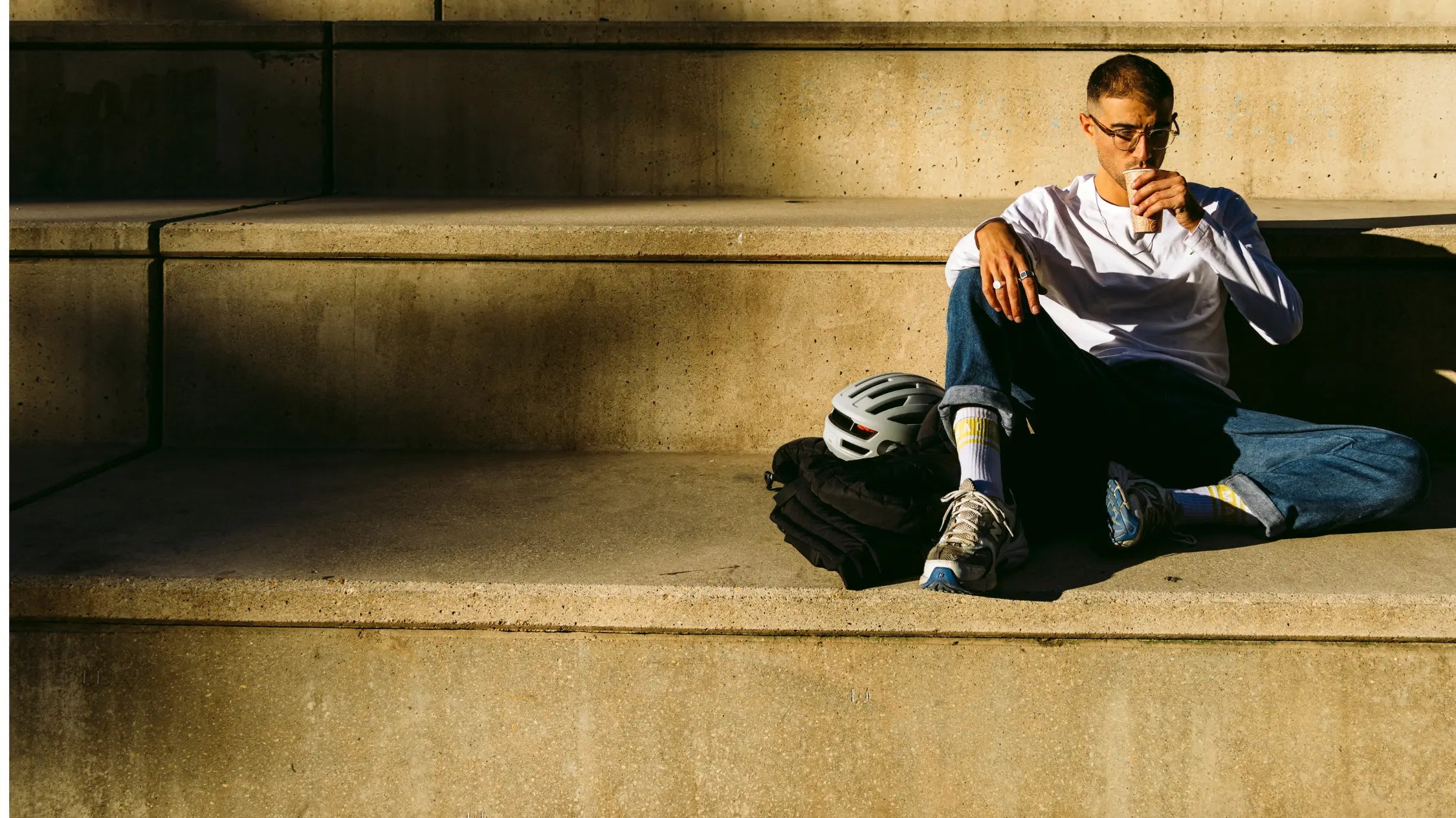 A person sits casually on concrete steps, sipping a drink. They wear glasses, a white long-sleeve shirt, jeans, and sneakers. A bike helmet and jacket are beside them, and sunlight casts shad