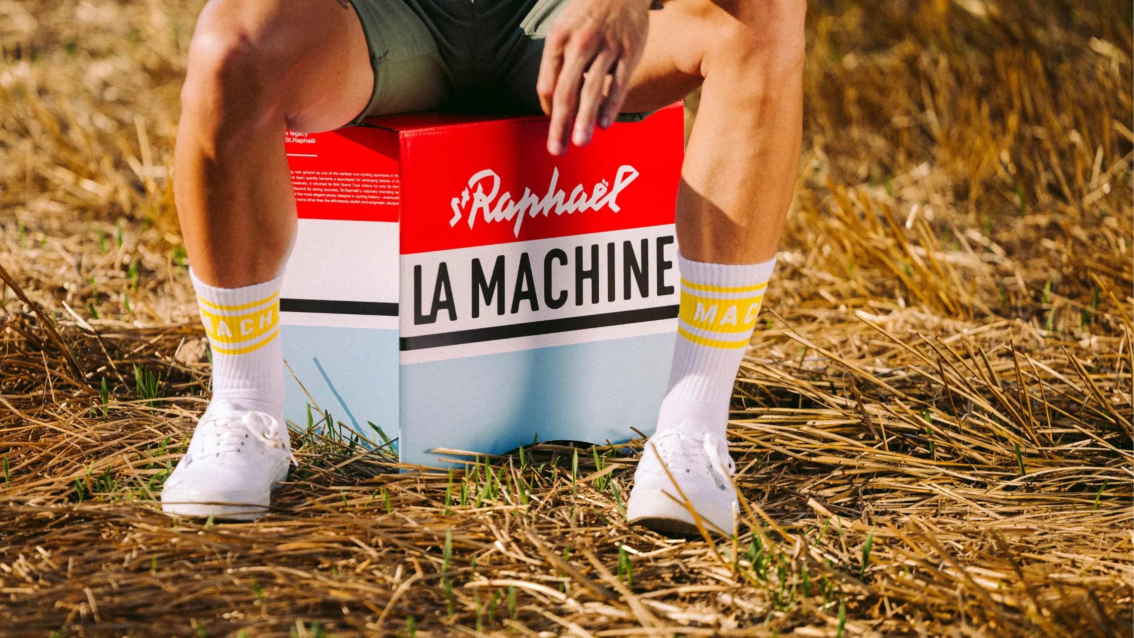 A person in white sneakers and yellow-banded socks sits on a red, white, and blue box labeled St. Raphaël LA MACHINE outdoors on dry grass. Their face is not visible.