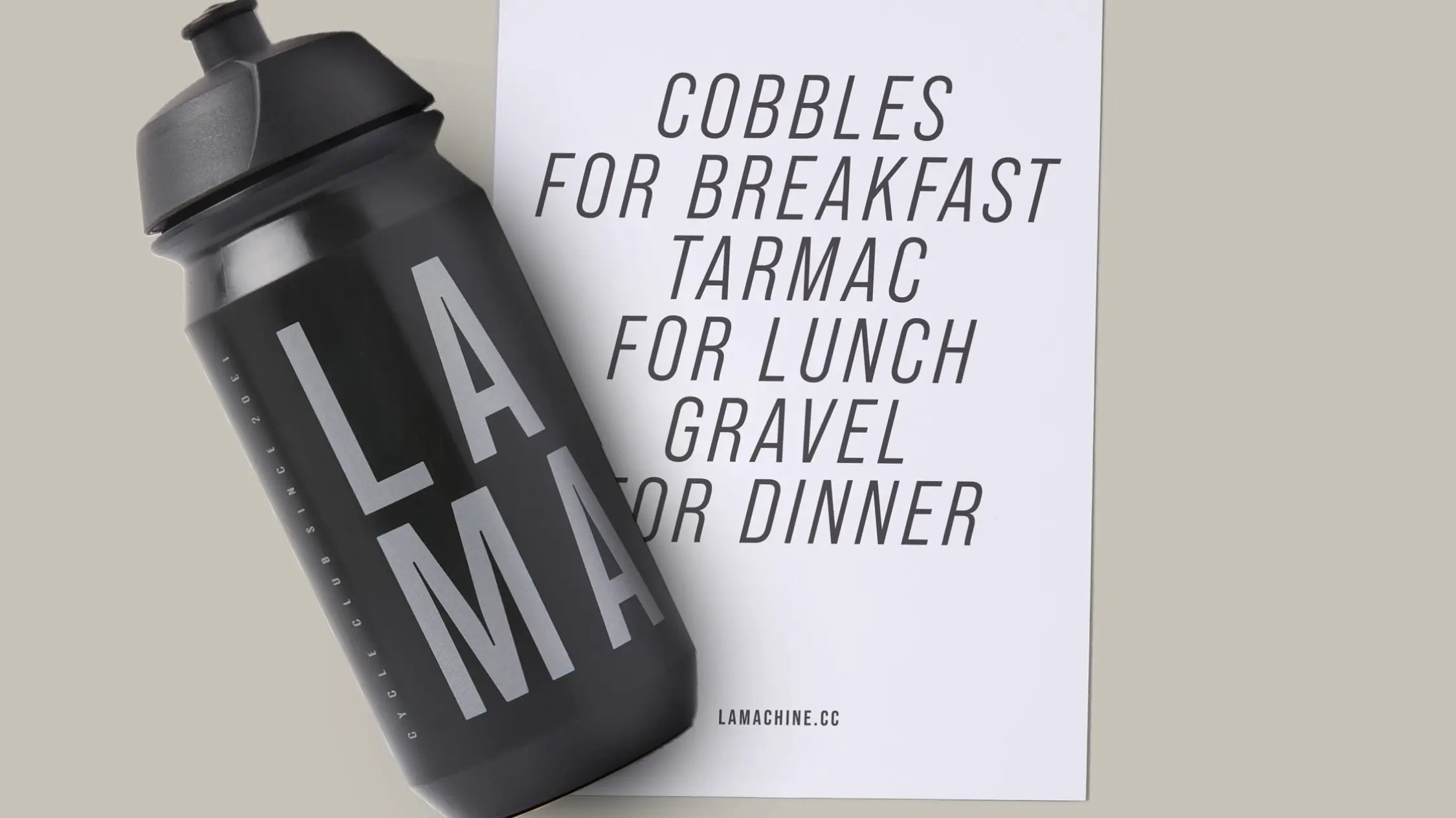 A black water bottle labeled LAMA lies next to a white card reading: Cobbles for breakfast, tarmac for lunch, gravel for dinner. Website lamachine.cc is at the bottom of the card.