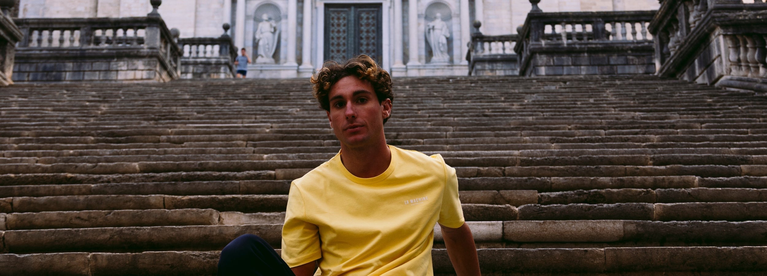A man in a grey hoodie, shorts, and a black cap lounges on a hay bale under a clear blue sky, wearing white socks with red writing and white sneakers.