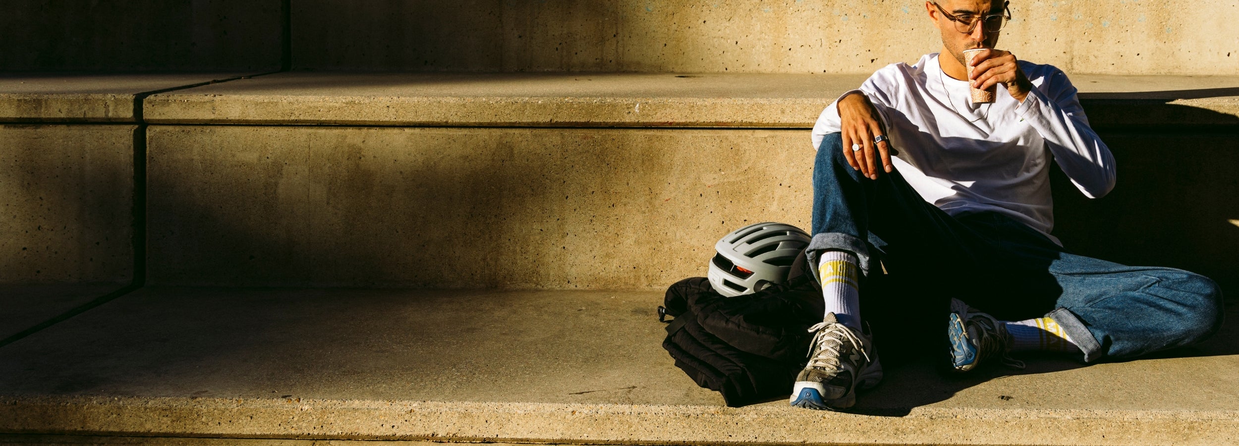 A person wearing glasses and casual clothes sits on concrete steps, sipping a drink. A bicycle helmet and dark jacket are placed beside them in the sunlight.