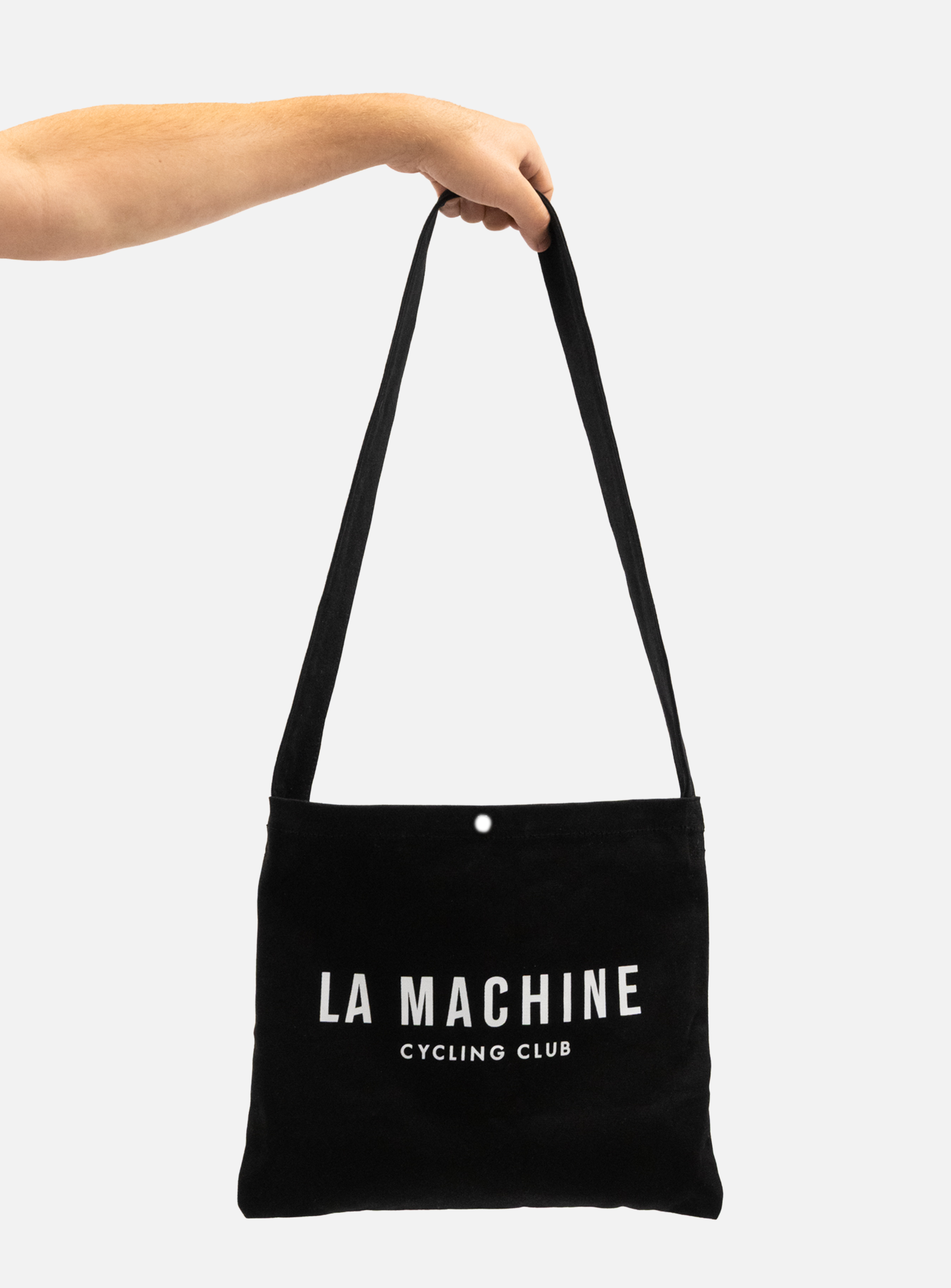 A hand holds the La Machine - Musette - Black from La Machine Cycle Club, featuring “LA MACHINE CYCLING CLUB” printed in white on the front, against a plain white background.