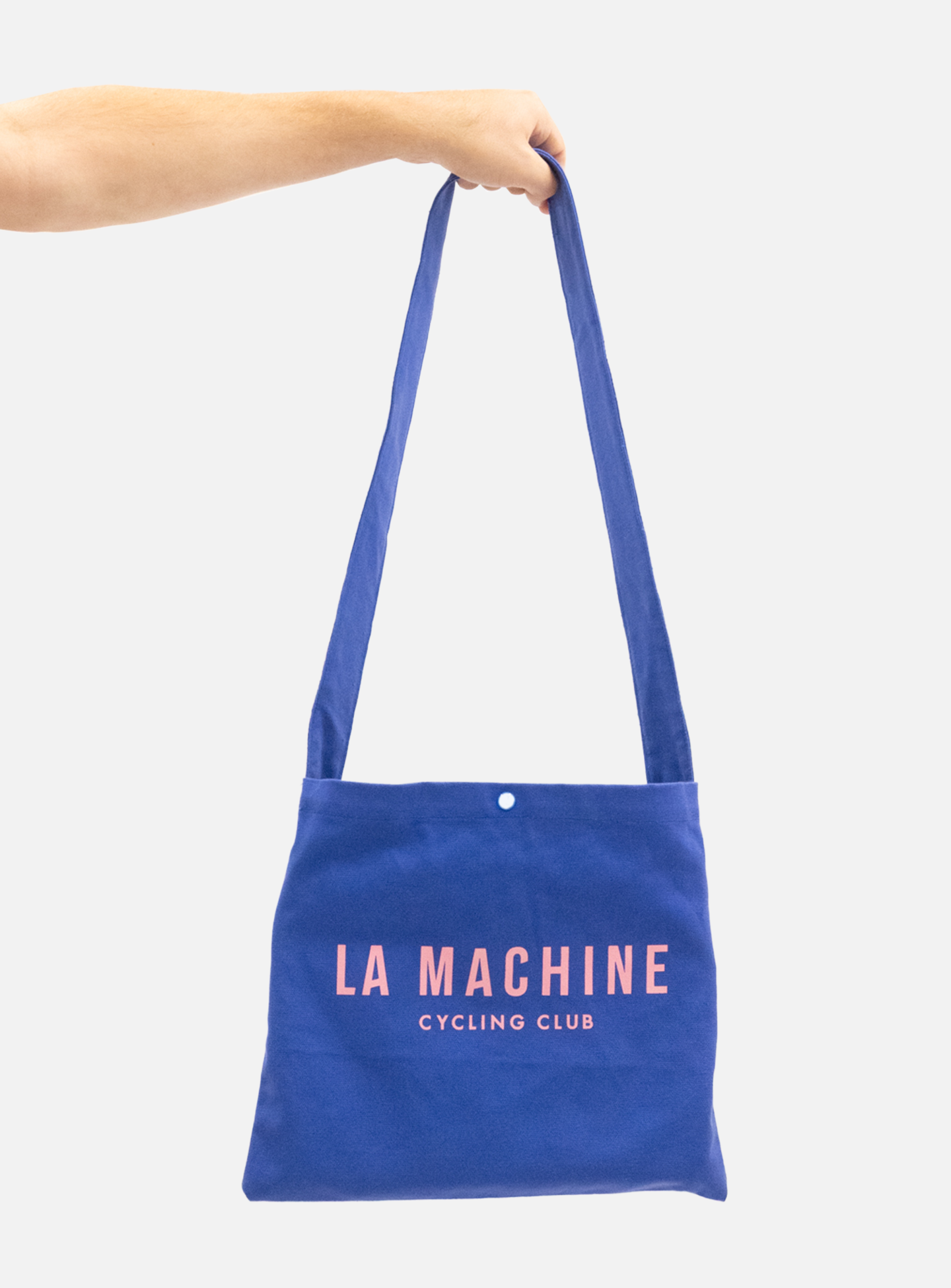 A hand holds up the La Machine - Musette - Blue by La Machine Cycle Club, featuring pink La Machine Cycle Club lettering on the front. The background is plain white.