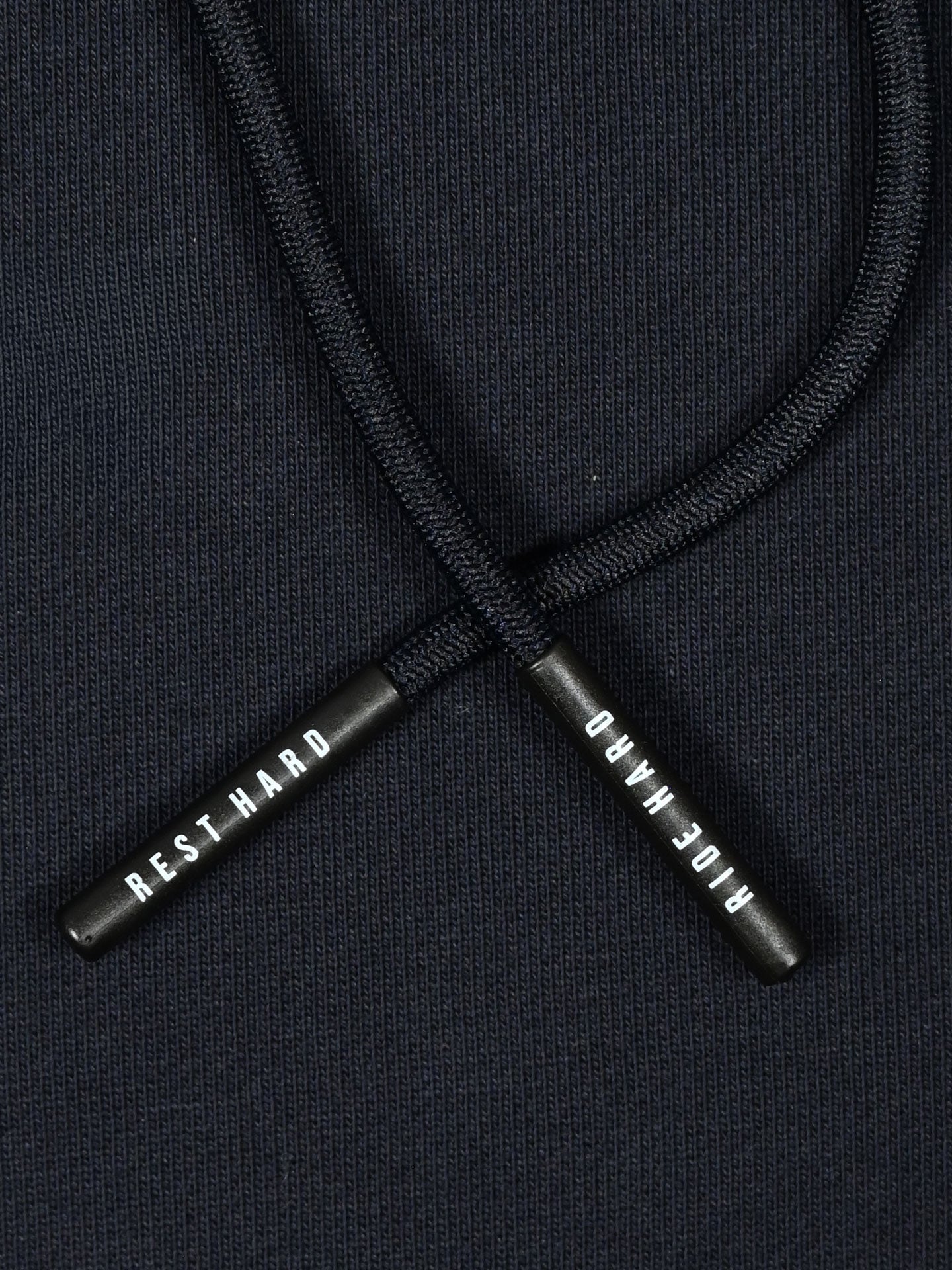 Close-up of two black drawstrings on the La Machine Cycle Club Ride Hard Rest Hard Hoodie; one tip reads REST HARD, the other RIDE HARD in bold white letters against dark 100% cotton fabric.