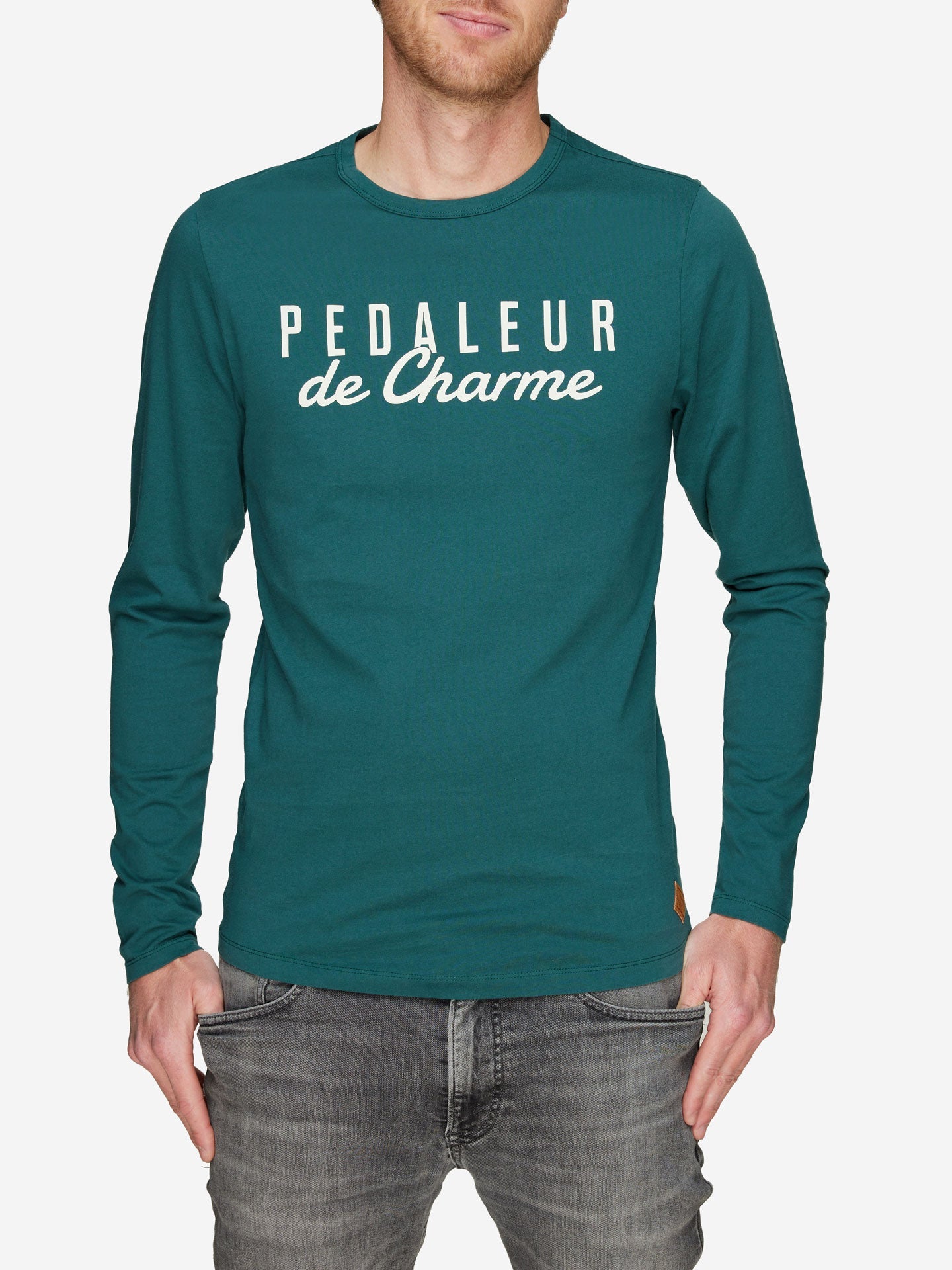 A man wearing a teal Pedaleur de Charme long sleeve t-shirt by La Machine Cycle Club, featuring white lettering, pairs it with gray jeans and stands against a plain white background.